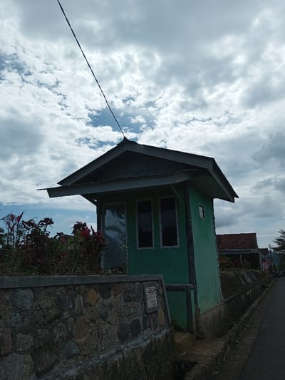 The scene depicts an outdoor environment in Ariyalai, Sri Lanka, under a sky characterized by scattered cumulus clouds and patches of blue. The primary subject is a small, single-story structure, approximately eight to ten feet in height, painted a light green color. It features a dark-colored gable roof with a corrugated metal overhang. An electrical wire extends diagonally upward from the apex of the roof towards the upper left. The building has two rectangular, dark-paned windows on its front-facing wall, and a smaller, similar window on its right-side wall. To the left of the prominent windows, a reflective, possibly metallic, panel or door is visible.
In the foreground, a low, robust stone wall, constructed from irregularly shaped, multi-colored rocks, spans the lower portion of the image. A flat concrete coping caps the wall, and a small, rectangular plaque is affixed to its lower right section. Behind this wall, to the left of the green building, a dense cluster of reddish-purple leafy plants and other green foliage is present.
Immediately in front of the stone wall and running parallel to it is a narrow concrete drainage ditch. Adjacent to the ditch, a paved road extends into the background, curving slightly to the right. In the distant midground, additional residential structures with reddish-brown tiled roofs are partially visible. No individuals, actions, or direct interactions are evident within the frame. The overall lighting is soft and diffused, consistent with an overcast or partly cloudy day.