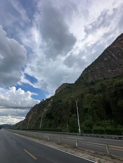 The image shows a multi-lane highway running alongside a steep, rocky mountainside covered in dense green vegetation. The sky above is dramatic, with a mixture of dark, heavy clouds and patches of bright blue sky. Several modern streetlights line the side of the highway, and power lines are visible crossing the frame in the distance. The highway itself is asphalt, with clear yellow and white lane markings. The mountainside dominates the right side of the image, with the rocky cliff face exposed in places, revealing layers of sediment and rock. The lower slopes are covered with a thick growth of trees and shrubs. In the far distance, the highway appears to curve into a more mountainous landscape. No people or vehicles are prominently visible in the scene.