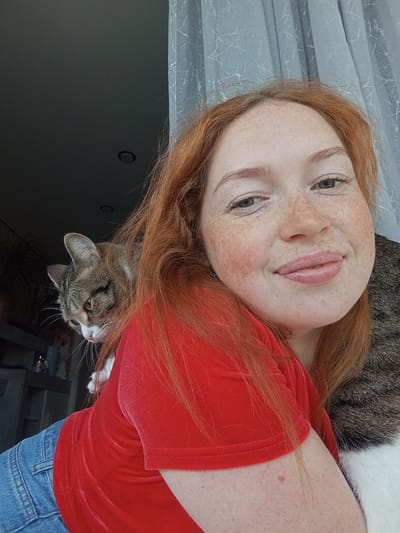 This is a horizontally oriented close-up photograph featuring a young woman and a domestic cat in an indoor setting. The woman, with fair skin and numerous freckles, has long, reddish-auburn hair. She is smiling directly at the camera, her lips slightly parted, revealing light pink lips. Her eyes are light-colored, possibly grey or blue. She is wearing a red top with a velvet or corduroy-like texture, and blue denim pants are partially visible in the upper left corner.
A tabby cat, with striped brown, grey, and white fur, is resting on the woman's upper back or shoulder. The cat's head is turned away from the camera, looking upwards and to the left of the frame, with its ears perked and whiskers visible. One of its white paws is also visible near the woman's neck.
The background is mostly dark and indistinct, suggesting an indoor environment. On the right side of the image, a light-colored curtain or blind with a subtle marbling or crackle pattern is visible. The lighting is soft and even, illuminating the subjects, suggesting daytime indoors. The overall mood is relaxed and affectionate. There is no visible text in the image. The location context for this image is Yurgamysh, Russia.
