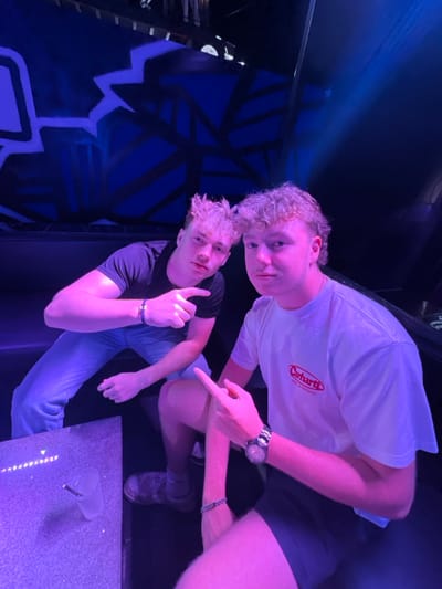 The image depicts two young adult males seated in a dark booth illuminated by prominent purple and blue ambient lighting, characteristic of a nightclub environment. The specific wall artwork, featuring abstract blue and black geometric shapes, identifies the location as Kiss Club in Albufeira, Portugal.

The individual on the left, with short, spiky blonde hair, wears a black short-sleeved t-shirt and light blue jeans. He is positioned leaning forward, extending his right arm with the index finger pointed directly towards the camera. A dark bracelet is visible on his left wrist.

The individual on the right, possessing curly blonde hair styled in a mullet, wears a white short-sleeved t-shirt with a red "Carhartt - Work In Progress" logo and dark shorts. He is also leaning forward, pointing his left index finger towards the camera. A metallic watch and a beaded bracelet are visible on his left wrist.

Both subjects maintain direct eye contact with the camera, indicating a posed photograph. In the foreground, on a speckled, reflective table surface, a clear plastic cup containing a beverage and a straw is visible. The scene suggests a social interaction captured in a nightlife setting.