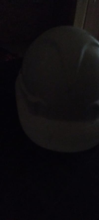 The image shows a dimly lit, close-up view of a white spherical object, possibly a ball or a portion of a larger object, against a dark background. The lighting is poor, with significant shadows obscuring details. There are faint indications of a seam or texture on the surface of the object. The environment appears to be indoors, with a vertical wooden structure visible in the upper right corner, suggesting a wall or furniture. There are no people or identifiable landmarks present. The image does not contain any specific information that would allow for its localization in Заречный ауылдық округі, Kazakhstan.