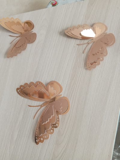 Three rose gold butterfly decorations are affixed to a light-colored wooden surface. The butterflies are decorative and appear to be made of a metallic material. They are positioned in a scattered arrangement across the surface. The top left butterfly is angled downwards and to the right, showing its patterned wings. The top right butterfly is facing towards the viewer and slightly upwards, with a strong reflection on its wings. The bottom butterfly is the largest and is positioned horizontally, extending from the left to the center of the frame, also displaying its detailed wings and body. The wooden surface has a visible grain pattern. No people or any other objects are present in the scene. This image was taken in Danilov, Russia.
