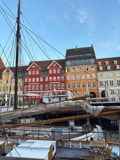 This is a vertically oriented, low-angle outdoor photograph of a canal scene in Copenhagen, Denmark. The foreground is dominated by the rigging and mast of a sailboat. The sails are furled and secured with ropes. A portion of the boat's deck is visible, featuring wooden structures and white bulkheads. In the background, a row of colorful, historic buildings lines the canal. The buildings are predominantly red, orange, and grey, with white-framed windows. The sky is a clear blue with a few wispy clouds. The lighting suggests it is daytime, likely morning or afternoon, with fair weather. The scene evokes a sense of historical charm and maritime activity. No people are clearly visible. Text is visible on a building in the background that reads "Hotel Bethel".