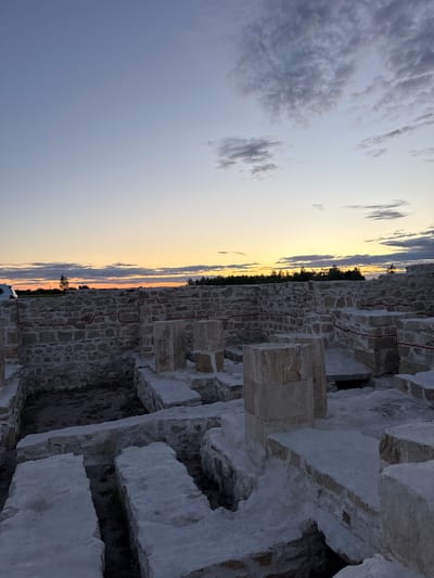 An archaeological site is shown at dusk. The ruins consist of stone walls and structures, some with concrete or plaster applied. Red wiring is visible along some of the stone walls. In the background, a line of trees forms the horizon, and the sky above transitions from a soft orange glow near the horizon to a pale blue and then a darker blue as it ascends. Scattered clouds are present in the upper sky. A portion of a white vehicle is visible on the left side of the image, partially obscured by the ruins. The scene appears to be part of the Bulgarian settlement ruins in Bolgar, Russia.