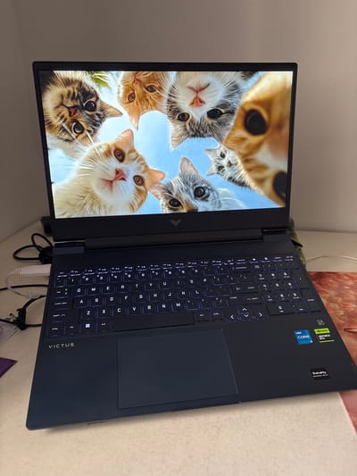 A black HP Victus laptop is open on a table. The screen displays a group of cats looking down at the viewer. The keyboard is visible with illuminated keys. Stickers indicating Intel Core i5 and Nvidia GeForce RTX are near the upper right corner of the keyboard area. The laptop is angled slightly to the right.