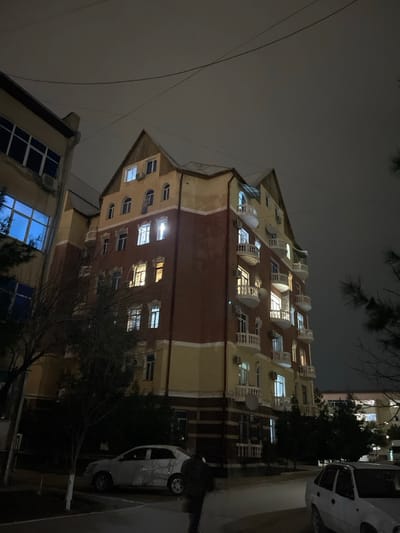 This nighttime photograph captures a street scene in Navoiy City, Uzbekistan. The central focus is a multi-story residential building, approximately six floors in height, featuring a gabled roof section and a facade segmented by color: a reddish-brown middle section flanked by lighter yellow or beige upper and lower portions, accented with white trim. Numerous windows are visible, many illuminated with either warm yellow or cool white light. The building displays multiple white, curved balconies and external air conditioning units, along with at least two white satellite dishes mounted on its exterior.

To the left of the central building, a section of an adjacent multi-story structure is visible, characterized by large, dark-paned windows, some reflecting a distinct blue light, and an external air conditioning unit. The sky above is dark and overcast, with several power lines traversing the upper frame.

At ground level, a paved area or sidewalk extends across the foreground. A lone individual, dressed in dark clothing, is observed walking away from the camera in the lower center-right of the frame. Two sedans are parked: a light-colored vehicle is positioned on the left, facing right, and a white sedan is partially visible on the far right, facing left. Sparse, bare-branched trees and some coniferous trees line the street and are situated near the buildings.