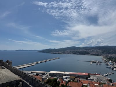 The image captures a panoramic view of the port and coastline of Kavala, Greece, from a high vantage point, likely from the city's fortress. In the foreground, a stone wall of ancient fortifications runs along the left side, with visible battlements and weathered stone. Beyond the wall, the deep blue sea stretches towards a crescent-shaped coastline lined with buildings.
A long concrete pier extends into the water, with several cargo containers stacked on it. Further into the harbor, a marina is filled with numerous yachts and smaller boats. The city of Kavala sprawls along the hills in the middle ground, a dense collection of white and light-colored buildings with red-tiled roofs. The background is dominated by lush green hills and mountains under a bright blue sky with scattered white clouds. The overall scene is one of a bustling port city integrated with historical structures and a beautiful natural landscape.