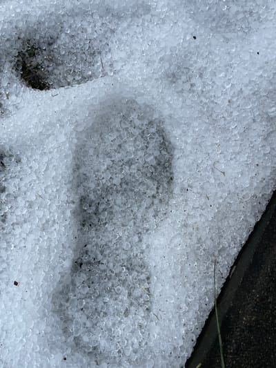 The image depicts a ground covered in hail, with a distinct footprint impression in the center. The hail consists of small, round ice pellets. The footprint appears to be from a shoe or boot, disrupting the uniform layer of hail. A dark, possibly concrete, edge is visible in the upper right corner. The background appears to be a mix of dirt and possibly some grass. The location, Olginka, Russia, is based on the prompt, but there is no specific indicator of location in the image.