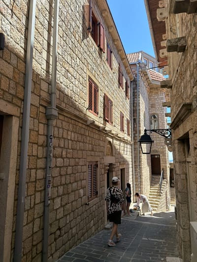 The image depicts a narrow, cobblestone street in the old town of Ulcinj, Montenegro. The street is lined with traditional stone buildings, characterized by their rough-hewn blocks and dark wooden shutters. A tall, grey downspout runs vertically along the left-hand building. In the mid-ground, a black wrought-iron lantern hangs from the side of a building. A set of stone stairs with a metal railing leads upwards to a wooden door.
Several individuals are present in the scene. In the foreground, a person wearing a black and white patterned shirt, dark shorts, a white cap, and sneakers walks away from the camera. Further down the street, two young women stand near the stairs. One is wearing a blue top and a dark skirt, while the other is wearing a light-colored outfit and appears to be bending down to retrieve an item. The bright sunlight casts sharp shadows across the street and buildings, suggesting a clear, sunny day. The sky is a vibrant blue. The overall impression is of a well-preserved historic urban environment.