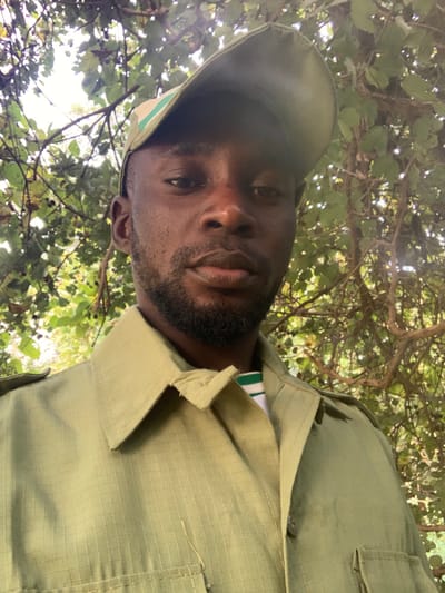A male individual is depicted in a close-up, outdoor portrait taken in Kubi A, Nigeria. He has dark skin, short dark hair, and a neatly trimmed beard, gazing directly forward with a neutral expression. He wears an olive-green, collared, long-sleeved shirt or jacket, possibly uniform, made of a textured ripstop-style fabric, with visible shoulder epaulets and indications of chest pockets. Underneath, a white t-shirt with green horizontal stripes around the collar is visible. He also wears an olive-green baseball cap with a lighter green stripe on the brim, positioned slightly askew. The background consists of dense green foliage, including leaves and branches, suggesting a natural, wooded environment. Bright, diffused light filters through the canopy from above.