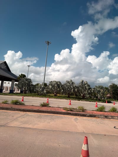 A low-angle medium shot captures a road in front of a white building with a covered walkway and a dark sloped roof, suggesting it might be a public facility or checkpoint. Several tall, slender palm trees with fan-shaped leaves line the area between the road and the building, interspersed with green grass and smaller bushes. A prominent tall light pole with multiple lamps stands near the center of the frame. The road is marked with white lines and lined with red and white traffic cones at intervals. The sky above is a bright blue, filled with dramatic, puffy white clouds. The foreground shows a concrete surface, possibly a sidewalk or a different lane of the road, with another red and white traffic cone in the lower right corner. The scene appears to be set in a tropical or subtropical environment.