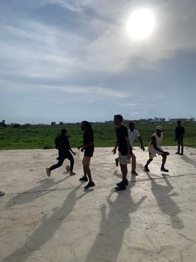 In Gwafan, Nigeria, a group of six young men are playing soccer on a concrete court. The sun is shining brightly in a partly cloudy sky, casting long shadows of the players onto the cracked concrete. The players are dressed in casual attire, including t-shirts, shorts, and athletic shoes. One player in the foreground is wearing a dark t-shirt and patterned shorts, while another, slightly behind him, is in a white short-sleeved shirt and light-colored pants. To the left, a player in a dark outfit is kicking a soccer ball. Further to the right, a player in a white tank top with yellow trim and white shorts is in motion, and another player in a dark t-shirt and tan shorts stands nearby. In the background, a man in a dark shirt and jeans stands to the far right. Beyond the court, a wide expanse of green grass stretches towards a horizon dotted with buildings and vegetation, suggesting a semi-urban or rural setting.