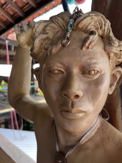 The image is a close-up, eye-level shot of a light brown, terracotta-like statue of a person's head and upper torso. The statue has curly hair with some weathered cracks and a metal loop with a chain attached to the top of its head. It appears to be outdoors, with a blurred background suggesting a rustic setting. The roof of a building with orange tiles is visible at the top of the frame, and what looks like corrugated metal roofing is present in the lower left. The statue's face is turned towards the camera, with its mouth slightly open and its eyes looking forward. A thin, light-colored cord is draped around its neck, with a round, dark pendant visible at the bottom center of the frame. The statue's right arm is raised behind its head, with its hand blurred and out of focus. The statue is located in Tejakula, Bali, Indonesia.