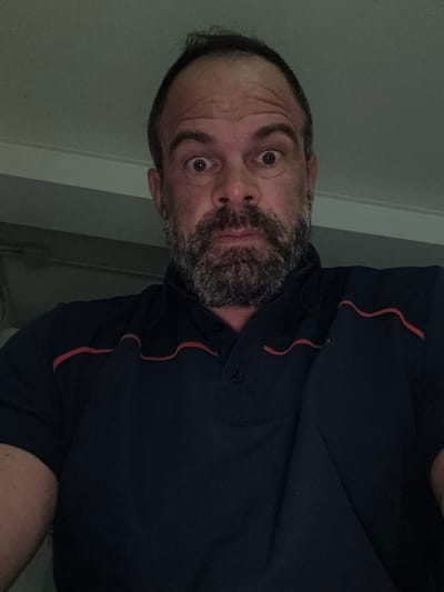 The original image, rotated ninety degrees clockwise, presents a close-up portrait of an adult man in what appears to be Diekirch, Luxembourg. The man has a dark beard with some graying, dark hair with a visible receding hairline, and is looking directly at the camera with wide, intense, or surprised eyes. His forehead shows wrinkles, suggesting a furrowed brow. He is wearing a dark, possibly navy blue, short-sleeved collared shirt. The shirt features distinctive wavy red trim details on the chest area. A button on his shirt is visible. The background is a plain, dimly lit wall, which appears light gray or greenish-gray, with subtle vertical lines that might indicate a doorframe or wall paneling. The low light suggests an indoor setting, possibly during the evening or night. There is no visible text in the image. The crops provide closer views of his eye and forehead, the texture of his beard, and the specific red wavy detailing on his shirt.