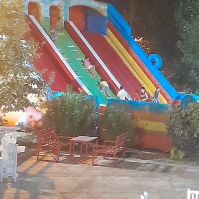 The image shows an inflatable slide with children playing on it. The slide is multi-colored with red, yellow, green, and blue sections. Several children are visible on the slide, some climbing, some sliding. In the foreground, there are red chairs and tables, and a white plastic chair. The setting appears to be outdoors, possibly in a park or recreational area, with trees and bushes visible in the background. The overall scene suggests a lively atmosphere of outdoor recreation. The location is identified as Rudəkənar, Azerbaijan.