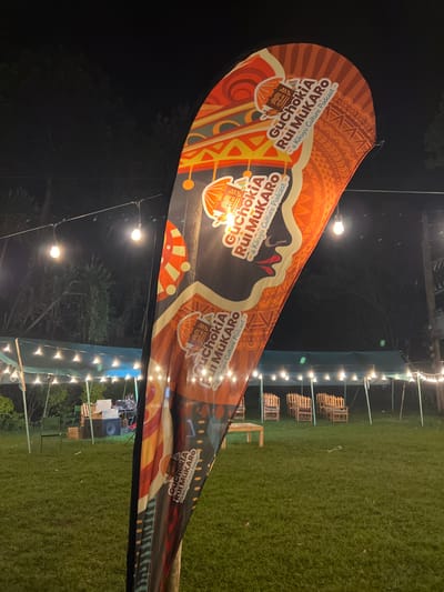 This media file captures an outdoor night scene in Mururiini, Kenya, featuring a large, brightly colored banner advertising a podcast. The banner, stretched across the foreground, displays traditional patterns in orange, red, and black, and prominently features the text "GuchokiA Rui MuKARO - a Kikuyu Culture Podcast -" repeated several times.
The setting appears to be prepared for an event or gathering, with string lights illuminating a grassy area in the background. Beneath these lights, several small wooden structures, resembling crates or pallets, are arranged, likely serving as seating or tables. Dark trees are visible behind the lighting setup, contributing to the nighttime ambiance. The sky is dark, indicating that the photograph was taken after dusk. The overall impression is one of an outdoor cultural event or social gathering. No people are visible in the image.