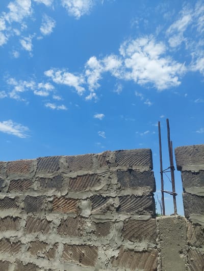 A low-angle, medium shot captures a section of a brick wall under construction against a bright blue sky filled with scattered white clouds. The wall, made of rough, unevenly mortared bricks, shows diagonal scoring patterns on many of the brick faces. Two vertical rebar rods protrude from the top of the wall, their tops bent and intertwined with a horizontal support rod. The background reveals a hazy glimpse of distant greenery and sky through a gap in the wall. The image appears to be taken outdoors, possibly in a construction site.