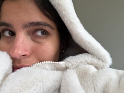 A close-up, eye-level image depicts the upper portion of an individual, likely an adult, in an indoor setting. The subject has dark hair and brown eyes, with visible dark eyebrows. Their gaze is directed slightly to their right (viewer's left). The individual wears a light-colored, likely off-white or cream, faux fur hooded garment. The hood is up and frames the upper right side of their head. The garment's zipper, featuring white plastic teeth, is partially visible and extends upwards, obscuring the subject's mouth and chin. The lower part of the face is covered by the zippered faux fur material. The background is a plain, light grey wall. The scene's specific location within Princeton, United States, is not discernible from the visual evidence.