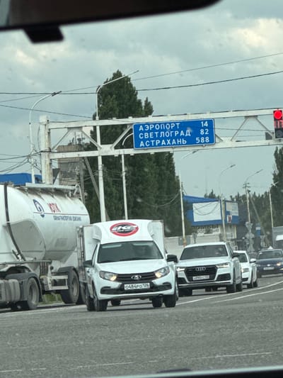 This image shows a street scene in Rozhdestvenskaya, Russia. The perspective is from inside a vehicle, looking out. The primary focus is a blue road sign indicating distances to various locations. The sign reads "АЭРОПОРТ" (Airport) with a distance of "14", "СВЕТЛОГРАД" (Svetlograd) with a distance of "82", and "АСТРАХАНЬ" (Astrakhan) with a distance of "588". The sign is mounted on a white metal structure, possibly an overpass or gantry.
In the background, there are several vehicles on a road, including cars and trucks. One white truck, possibly a cement mixer, is prominent on the right side of the frame. Another white vehicle, an SUV, is also visible. The sky is overcast, suggesting a cloudy day. Green trees line the side of the road in the distance. A traffic light with a red signal is visible at the top left of the image. The overall scene appears to be during daylight hours. The road surface is asphalt. There is no clear indication of people within the vehicles. The image has a slightly grainy quality, and the overall lighting is diffused due to the cloud cover.