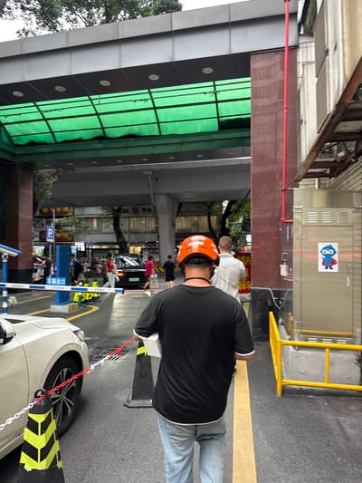 A man wearing an orange safety helmet and a black t-shirt walks away from the camera, carrying a white box. He is in an outdoor urban setting, likely a parking lot or street. In the background, a white car is parked, and other people are visible walking in the vicinity. There are several traffic cones with yellow and black stripes, and a chain barrier. The environment appears to be covered by a green and grey structure overhead. The scene is set in Yuexiu District, China.