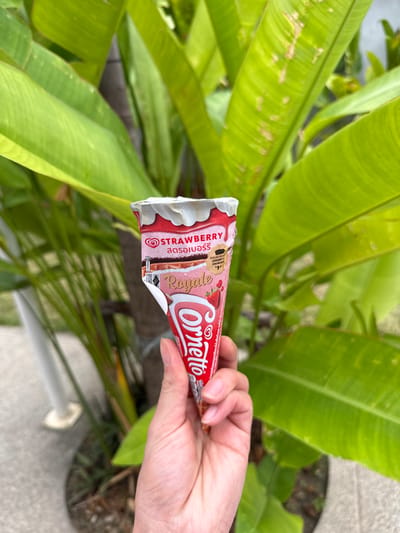 A light-skinned human hand holds a partially unwrapped Cornetto Royale Strawberry ice cream cone. The cone's wrapper is red and white, displaying "STRAWBERRY" in English and Thai script, along with "Royale." The white ice cream is visible at the top of the cone. The background consists of dense, broad green tropical plant leaves, with a dark tree trunk partially visible between the foliage. A grey paved surface and a white cylindrical object with a circular base are visible in the lower left background. The scene is set in Kamala, Thailand.