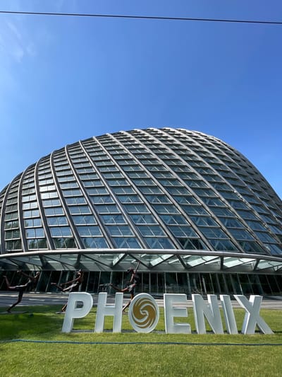 The image shows the Phoenix International Media Center in the Chaoyang District of Beijing, China. The building is a curved, metal-clad structure with numerous rectangular windows. In front of the building is a large sign that spells out "PHOENIX", complete with the company's logo. Three statues of people performing various activities are positioned adjacent to the sign and the base of the building. The surrounding area consists of green grass and a clear blue sky.