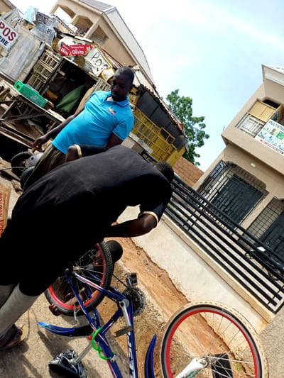 In Garko, Nigeria, two men are near a blue bicycle. One man, wearing a blue polo shirt, stands with his head lowered. The other man, dressed in a black shirt and grey pants, is bent over, working on the bicycle. The bicycle is blue with red accents on its wheels and has a green lock on its frame. The scene appears to be in an urban outdoor setting, with buildings and market stalls visible in the background. The ground is paved with dirt and concrete. Signs are present in the background, one of which reads "POS".