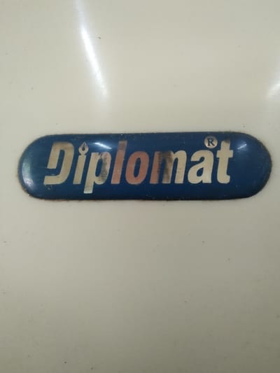 The image shows a close-up of a blue oval sticker with the word "Diplomat" in stylized silver and orange letters. The sticker is affixed to a light-colored, possibly white, surface. A small registered trademark symbol "®" is visible above the letter "t". The sticker appears to be slightly worn. The background is uniformly light-colored, showing subtle variations in lighting and some minor dust specks. There is no indication of the city of Tervel, Bulgaria, in the image.