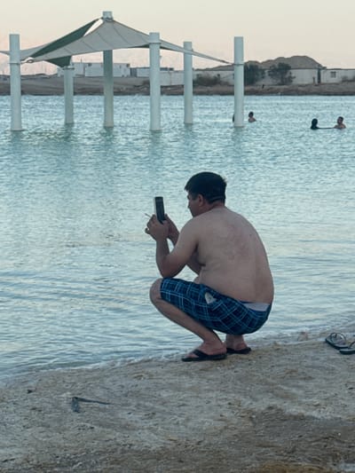 The image shows a man squatting on the sandy shore of a body of water, possibly the Dead Sea, in Tamar Regional Council, Israel. He is holding a smartphone and a cigarette. The water appears to be still and slightly rippled, with some structures and people visible in the background. The sky is overcast and the overall lighting suggests it is late afternoon or early evening. The man is wearing plaid shorts and flip-flops.