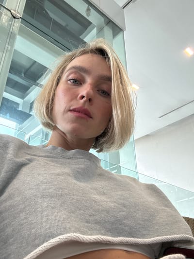 A blonde woman with light skin, blue eyes, and a slight smirk is looking up at the camera. She has on a grey sweatshirt and her hair is cut into a bob. The background is a blurry, modern interior with large windows and bright overhead lighting, suggesting an indoor public space such as an airport lounge or waiting area. The lighting is bright and diffuse, casting soft shadows on the woman's face and clothing. The scene is well-lit and the focus is on the woman's face, with the background out of focus. No specific location in Mai Khao, Thailand is identifiable from the visual elements of the image.