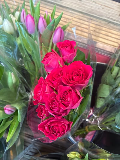 A close-up shot shows several bunches of flowers wrapped in clear plastic sleeves. Predominantly, red roses are in focus, with pink tulips also visible in the foreground. The flowers appear to be displayed on a light brown, wooden surface, likely a table or shelf. The background hints at other flower arrangements, with green stems secured with white ties. The setting appears to be a florist shop or market stall. The location is Oswestry, United Kingdom.