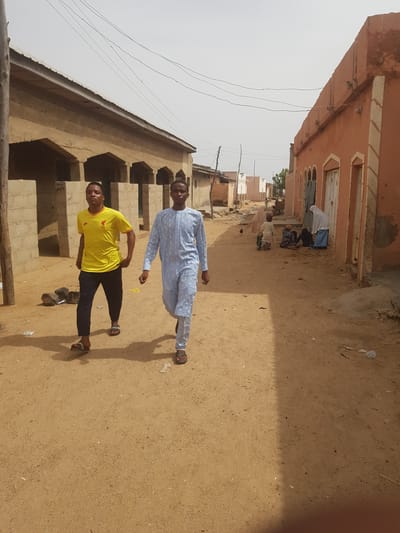 Two young men stand in a dusty, sunlit street in Dutsinma B, Nigeria. One wears a yellow t-shirt and dark pants, while the other is dressed in a light blue robe. The street is lined with buildings of earthen construction. In the distance, several individuals are seen praying near a building with peach-colored walls. Power lines extend across the sky.