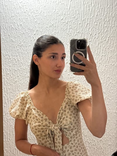 A woman is taking a selfie in front of a textured white wall. She is wearing a light-colored floral dress. She is holding a black smartphone with a clear case. The phone has a MagSafe ring visible on the back. She is facing the camera with a slight tilt of her head. The location is not evident from the image, and there is no indication of being in Miljevci, Montenegro.