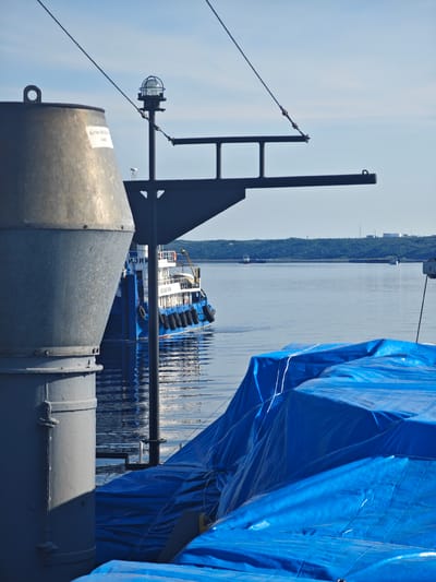 A bright, sunny day at a Russian port. In the foreground, a large, cylindrical metal object dominates the left side of the frame, partially obscuring a blue and white ferry. The ferry, adorned with black fenders, is docked in the calm, reflective water. To the right, several large objects are covered in vibrant blue tarps, suggesting cargo or equipment being protected from the elements.

In the background, a chain of green, tree-covered hills rises from the horizon. Distant industrial structures, including tall smokestacks and storage tanks, are visible on the far bank, hinting at the port's commercial activity. The sky is a clear, pale blue, with only a few wispy clouds.

The scene is peaceful, with the gentle lapping of water against the ships. A narrow black pole with a light fixture stands tall in the middle ground, casting a faint shadow. The overall impression is one of a functional, working port on a pleasant day. There is some visible text on a white label on the large metal cylinder in the foreground.