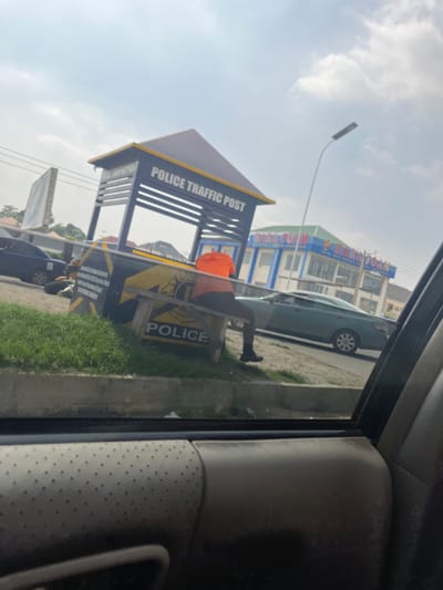 The image depicts a street scene in Dutsen Alhaji, Nigeria, viewed from inside a vehicle. The primary focus is a police traffic post with a distinctive yellow and dark blue design. The post has a roof and open sides with railings, and text on the side reads "POLICE TRAFFIC POST." Inside the post, a person wearing an orange shirt is visible.
In the background, across the street, is a multi-story building with the prominent text "HANA FOAM" in blue and green letters. A light-colored car is parked in front of this building. The sky is overcast with clouds, suggesting it is daytime, likely morning or afternoon. The area appears to be a commercial or urban setting, indicated by the building and the presence of the traffic post. The overall atmosphere is ordinary, capturing a moment of daily life in the depicted location. The image is taken with a slight tilt, giving it a dynamic perspective.