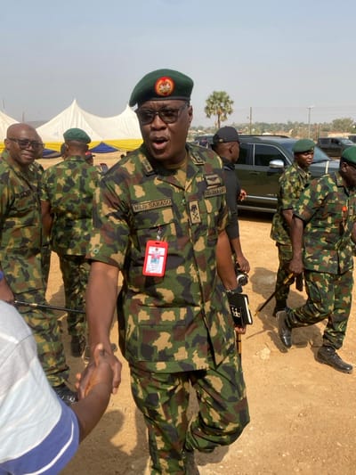 This image captures an outdoor scene in Kachia, Nigeria, under bright daylight with a clear, hazy sky. The ground is unpaved, consisting of light brown dirt or sand.
In the foreground, a dark-skinned male individual, central to the frame, is depicted wearing a Nigerian Army camouflage uniform, a green beret adorned with a national emblem, and black-rimmed glasses. His uniform displays a name tag "M.L.D. SARASO" on the left chest and a red identification badge suspended from his neck. Rank insignia on his right shoulder appears to include three stars and an eagle emblem, with "NIGERIAN ARMY" text above it. His mouth is open, suggesting speech or a momentary expression. His right hand is extended, shaking the hand of an individual partially visible at the bottom left, wearing a blue and white striped shirt. In his left hand, he holds a black notebook and a dark object, possibly a pen.
Several other male individuals in Nigerian Army camouflage uniforms and green berets are visible in the background and mid-ground. One uniformed man with glasses is smiling to the left of the central figure. Further back, other uniformed personnel are seen standing and walking; one individual to the far right carries a dark stick or cane. A non-military individual in a black shirt and cap is partially observed behind the central figure.
The background features several large white tents with yellow bordering, indicative of an organized outdoor event. A dark-colored SUV is parked further back on the right side of the frame. Distant palm trees and low-lying hills or elevated terrain are visible against the horizon.