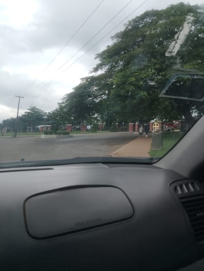 The image, captured from inside a vehicle, looks out onto a street scene in Iju, Nigeria. The sky is overcast and grey, with power lines running across the top of the frame. On the left side of the street, there are several large trees and what appears to be a manicured lawn. Further down the street, a building with red brick pillars and white structures is visible. On the right side of the street, a large tree with dense green foliage dominates the view. A person, a woman, is walking on the sidewalk, carrying a bag. Beside her, another person, a man, is walking in the same direction. Behind them, a sign with "GRAILLAND" written on it is attached to a building. A gold circular emblem is also visible on the building. A trash can is situated on the sidewalk. The dashboard of the car is in the foreground, with an "AIRBAG" warning visible on the passenger side.