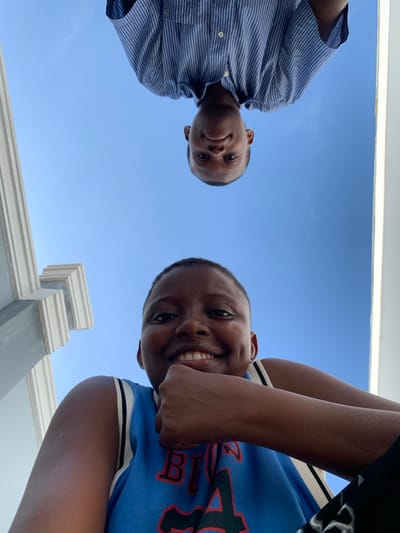 This is a low-angle portrait photograph featuring two young individuals looking downwards directly at the camera, set against a clear blue sky. The image was taken outdoors, likely in Ibasa, Nigeria, during daylight hours under sunny conditions.

On the left side of the frame, a person with dark skin and short dark hair is wearing a blue and white striped collared shirt. They have a subtle, content smile and are looking towards the camera. On the right, another person, also with dark skin and short dark hair, is smiling broadly, showing their teeth, and appears to be very happy. This individual is wearing a blue basketball-style jersey with black and white striped trim and partially visible red and black lettering on the front. Their arms are bent, with their forearms and hands partially visible.

The majority of the background is a bright, cloudless blue sky. At the very top and bottom edges of the image, portions of a light-colored architectural structure, possibly a building facade, a ledge, or a railing, are visible, suggesting the subjects are positioned above the camera. The overall mood of the image is joyful and candid, capturing a moment of casual interaction with the photographer. The text on the right person's jersey is not fully discernible.