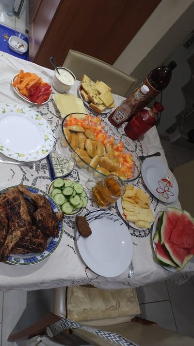 A dining table is laden with a spread of food, suggesting a meal or gathering. Various dishes are arranged, including grilled meats, a platter of small pastries, sliced watermelon, cut cheese, cucumber slices, and a bowl of chopped bell peppers and carrots. There are also bottles of ketchup and a dark liquid, a glass of amber liquid, a bowl of dip, and several plates. A slice of dark bread sits on a plate, and a piece of cheese is visible. The setting appears to be indoors, possibly a dining room, with a tablecloth covering the table. The lighting suggests it might be daytime or well-lit indoors. The overall scene depicts a generous offering of food ready to be enjoyed. No people are visible in the image. Visible text includes "HEINZ TOMATO KETCHUP".