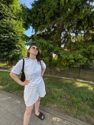 A young woman stands on a paved walkway, her body angled towards the camera. She is wearing a white t-shirt and white shorts, and black slip-on sandals. A black crossbody bag is slung over her left shoulder. She has long brown hair and is wearing sunglasses. Her left hand rests on her hip, and her right hand is placed on her right thigh. The woman appears to be posing for the photo.
The setting is outdoors, with a grassy area and trees visible in the background. Sunlight is casting shadows, suggesting it is daytime, likely late afternoon given the angle of the shadows. A chain-link fence is partially visible along the edge of the grassy area. The location context is Slonim, Belarus. There is no visible text in the image.