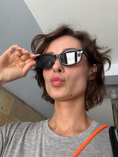 A young woman with short, wavy brown hair is shown from the chest up, wearing black sunglasses and a grey t-shirt. She is holding the left side of her sunglasses with her right hand and puckering her lips, creating a "duck face." The background consists of a textured grey wall and a tiled wall. An orange strap of a bag is visible over her right shoulder. The lighting suggests an indoor setting, possibly a hallway or residential interior, with a faint reflection in her sunglasses indicating an external light source. There is no indication of the city of Kiel, Germany.