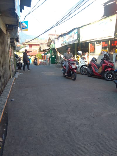The image shows a street scene in Cisarua, Indonesia, on a sunny day. The street is paved with asphalt and has a visible texture of parallel grooves. Several motorcycles are parked or being ridden on the street.
In the foreground, a man is riding a red scooter. He is wearing a grey t-shirt and dark pants. To his right, a white scooter is parked, and beyond that, a red motorcycle is also parked. Another red motorcycle, with a rider wearing a helmet, is further down the street on the right. A man in a dark jacket and pants is standing near the left side of the street, facing away from the camera and towards the parked motorcycles.
The buildings lining the street are typical of a commercial area. On the left, a building with a corrugated metal fence is visible. On the right, a row of shops with awnings are present. One shop has signage indicating "MIE BASO SOTO MIE BOGOR" and another displays "MIE AYAM". Electrical wires crisscross overhead. In the background, buildings with red roofs and some greenery are visible, suggesting a hilly or mountainous terrain, consistent with Cisarua's location. The sky is bright with a slight haze. Several pieces of litter are scattered on the street and sidewalk.