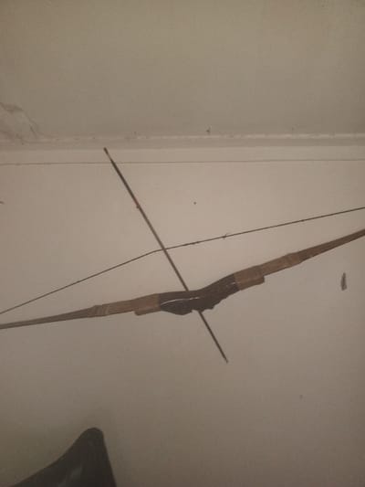 A bow and arrow hang against a white wall. The bow, made of wood, is dark in the center with lighter brown tips. The string stretches tautly between the bow's ends. An arrow intersects the bow's center, pointing downwards. A dark, indistinct object is partially visible in the lower left corner. The wall has minor imperfections near the ceiling.