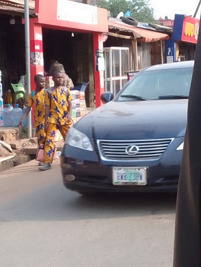 The image displays a street scene in Nkwelle Ezunaka, Nigeria, captured from a low angle, likely from within a vehicle. In the foreground, the front of a dark blue Lexus sedan occupies the right and center, featuring its grille, Lexus emblem, headlights, and a Nigerian license plate registered as "BWR-23BW".
To the left of the sedan, two young males, dressed in identical yellow outfits with blue patterns, walk along a dirt shoulder adjacent to the asphalt road. The male on the left carries a pink bag, while the male on the right has a light-colored strap across his right shoulder.
In the background, a red-painted storefront with an open entrance is visible. Outside this shop, large bottles of water and shelves displaying various beverages are present. Inside the entrance, a person wearing patterned clothing is seated. Adjacent to the red storefront on the right, another structure features corrugated metal siding, an awning, and two discarded tires on its roof. A glass display case and a sign bearing the partial text "VICTOR" are also visible. Further to the right, a blue sign with yellow text is partially obscured. The scene is illuminated by daylight.