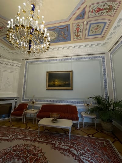 The photograph depicts the interior of a grand room, likely a parlor or salon, located in the Mikhailovsky (or Constantine) Palace in Strelna, Russia. The room is lavishly decorated, featuring a large crystal chandelier hanging from a painted ceiling adorned with classical motifs and intricate geometric patterns. The walls are a pale blue, bordered by a decorative frieze with a repeating circular pattern in blue and white.
Dominating the wall opposite the viewer is a landscape painting in a golden frame, depicting a dramatic scene with a ship at sea under a twilight sky. Below the painting, a red velvet sofa with tufted upholstery is placed between two small white side tables. To the left of the sofa, a matching red velvet armchair is positioned next to a white fireplace with ornate detailing. Another small white side table stands between the armchair and the sofa. On the sofa, a decorative bowl sits on a rectangular coffee table. To the right of the sofa, a large potted palm plant adds a touch of greenery. The floor is covered by a large, ornate rug with a predominantly red and beige pattern. The overall impression is one of opulence and historical significance.