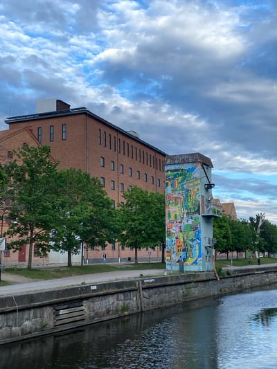 This image, set in Uppsala, Sweden, captures a tranquil urban waterside scene dominated by a large, historic building. The main subject is a tall, multi-story brick building with a dark roof and numerous small, rectangular windows evenly distributed across its facade.
A striking feature of the building is a large, colorful mural covering a significant portion of one of its end walls. This vibrant artwork depicts a whimsical and detailed landscape or town scene in a cartoonish style, featuring a variety of colorful houses, buildings, trees, hills, and possibly abstract shapes or figures. The mural's bright palette stands out against the muted tones of the brickwork and the surrounding environment.
To the right of the building, a calm body of water, likely a canal or river, flows. Its surface reflects the sky above, showing a mix of light and shadow. A sturdy stone and concrete embankment, with visible texture and some greenery, separates the water from a narrow path or strip of land. Several mature trees with lush green foliage are interspersed along this area, partially obscuring the lower parts of the building and lining the waterfront.
The sky is partly cloudy, with patches of clear blue visible between fluffy white and grey clouds. The overall lighting suggests either late afternoon or early evening, creating soft illumination across the scene. No specific activities are ongoing, and no prominent people are visible. No discernible text is readable within the image.