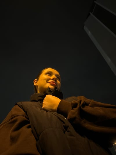 A low-angle, night-time photo depicts a person looking up. The subject is a fair-skinned individual wearing a dark jacket over a brown hooded sweatshirt. The background is dark, suggestive of night, with artificial lighting visible in the upper left corner. The location is potentially Akulovo, Gp Kubinka, Russia, based on the user's prompt.