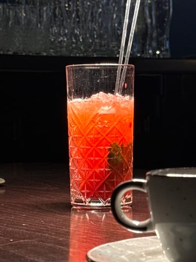 The image presents a brightly lit, close-up view of a cocktail in a patterned glass. The cocktail is a vibrant red color and appears to be garnished with a small green sprig. Two clear straws protrude from the drink. The glass sits on a wooden surface with a coffee cup in the upper right corner of the frame. The background is dark, with a textured panel visible behind the drink. There is no information in the image to identify its location in the city of Kursk, Russia.