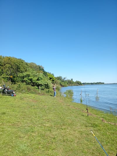 The image shows a scene by the shore of a body of water, likely a river, in Corrientes, Argentina. The sky is clear and blue. In the middle ground, a man is standing near the water's edge, holding a fishing rod. Another fishing rod is stuck in the grass near the water. To the left, there's a motorcycle parked on the grassy bank. The shoreline is lined with grass and some vegetation, bordering the water. Behind the motorcycle, there is dense green foliage forming a tree line. The water is relatively calm, with a few wooden posts visible.