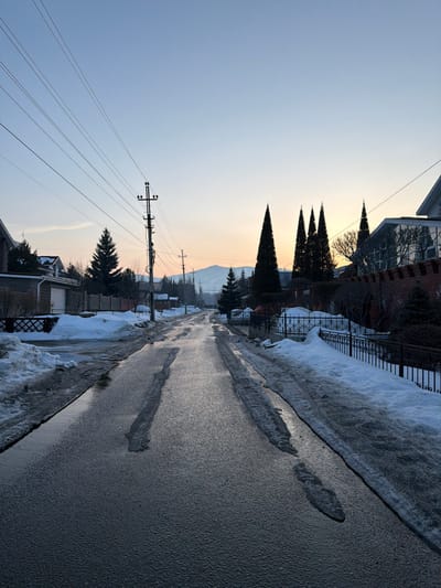 The image depicts a residential street in Zelenaya Polyana, Russia, during what appears to be either sunrise or sunset, characterized by a clear sky transitioning from light blue overhead to hues of orange and yellow at the horizon. The asphalt road, wet and partially covered with slush and melted ice, shows two distinct parallel paths of clearer asphalt, suggesting recent vehicle traffic. Snowdrifts and slush piles line both sides of the road.
On the left side, a short, dark fence precedes a house with a dark roof and a light-colored garage door. Several coniferous trees, including a prominent dark green one, are visible. Multiple utility poles, featuring horizontal crossbars and numerous overhead electrical wires, run along this side of the street, receding into the distance.
On the right side, an ornate black wrought-iron fence runs parallel to the road, backed by a red brick wall. Behind this wall and fence, a cluster of tall, slender, dark coniferous trees (likely cypress or similar species) stands prominently, silhouetted against the horizon. A modern house with reflective windows is partially visible behind these trees and the wall.
In the far background, snow-covered mountains or hills are visible, silhouetted against the pale sky. No individuals, animals, or active vehicles are present in the scene. The lighting suggests a low sun, casting reflections on the wet road surface.